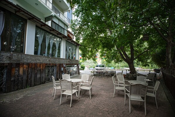 An outdoor seating area with wicker chairs and tables, a building with large windows, and a tree providing shade.