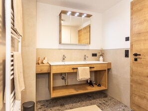 Ein Badezimmer mit einem Holzkosmetikschrank, zwei Waschbecken, einem Spiegel mit Beleuchtung, einem Handtuchhalter und einer Mülleimer.