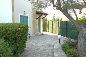 Un camí de pedra que condueix a un edifici amb una porta i persianes verdes, envoltat de vegetació i una tanca.