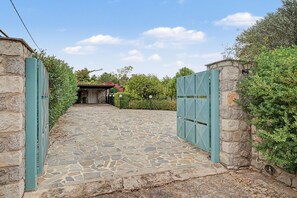 A stone-paved entrance with a metal gate, a stone wall, and a building with a tiled roof in the background. - Welcome to Villa Almira: Detached 3-Bedroom Retreat with Private Pool (Κρανίδι)