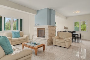 A living room with a fireplace, a sofa, a chair, and a dining area with a table and chairs. - Welcome to Villa Almira: Detached 3-Bedroom Retreat with Private Pool (Κρανίδι)