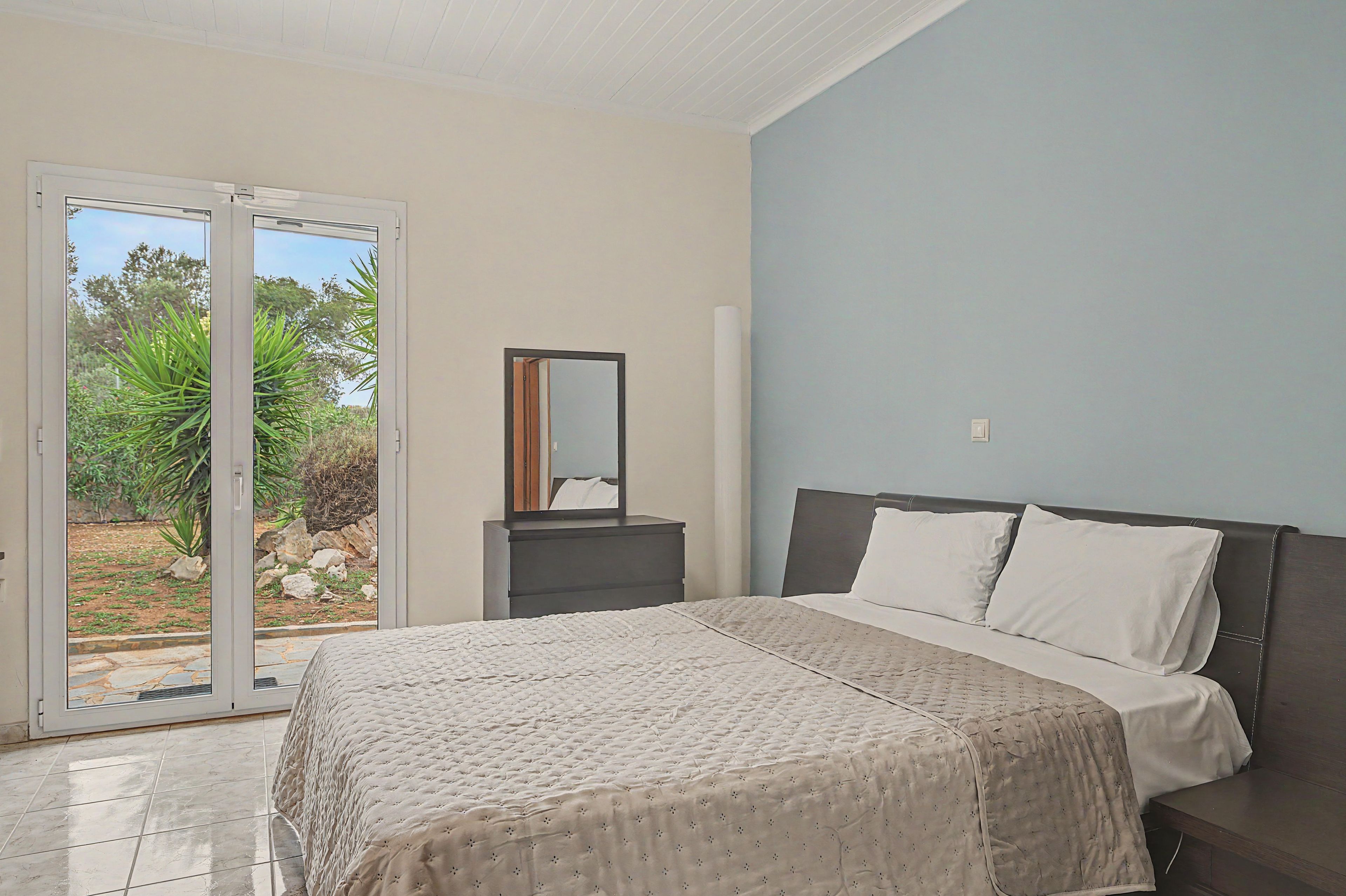 A bedroom with a bed, a nightstand, a mirror, and a sliding glass door leading to a garden.