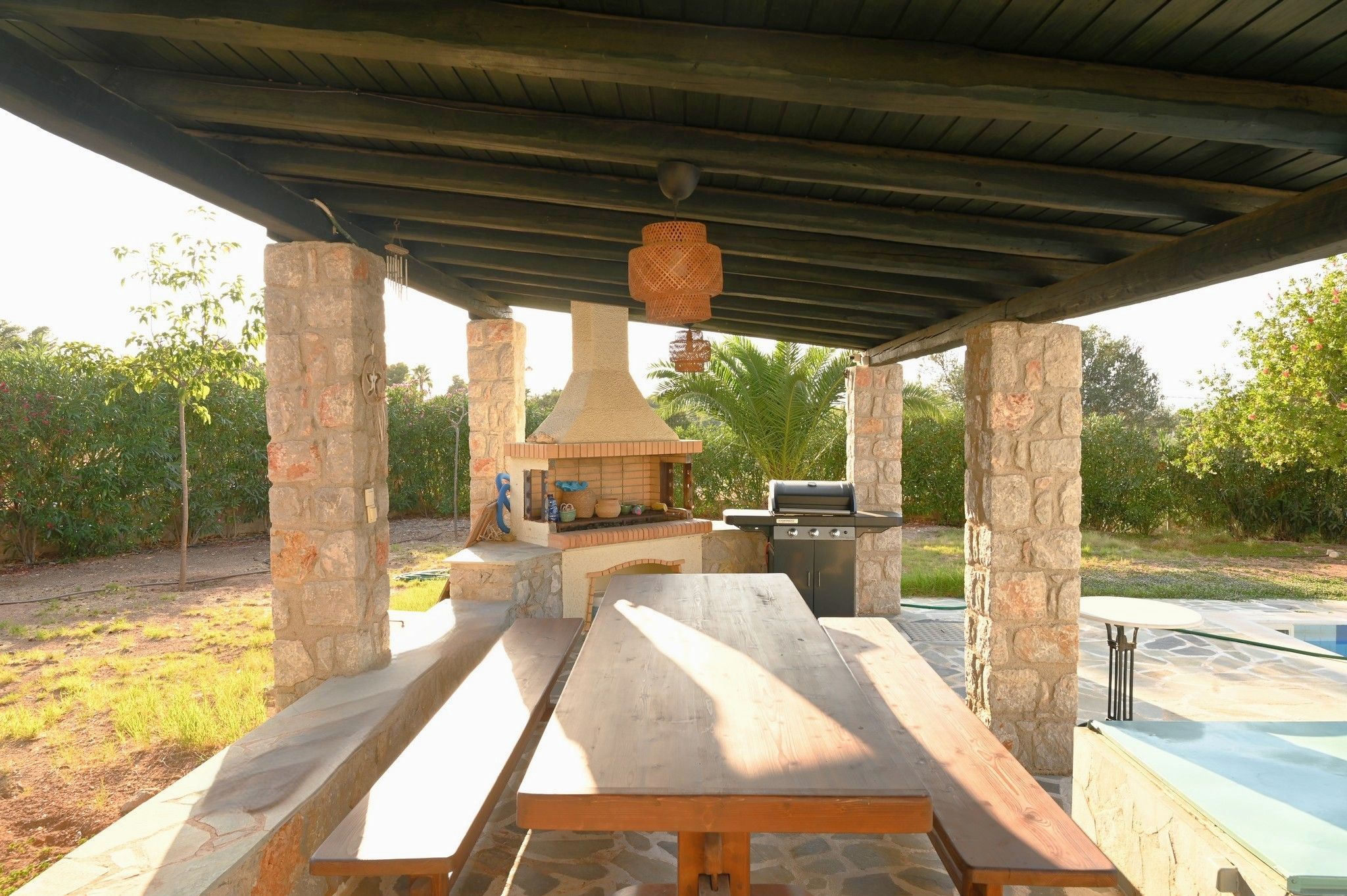An outdoor patio with a stone barbecue and grill area, surrounded by stone pillars and a covered roof.