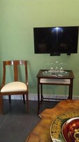 Una stanza con televisore a parete, una sedia di legno con cuscino, un tavolino con piano in vetro e un piatto decorativo su un tavolo di legno.
