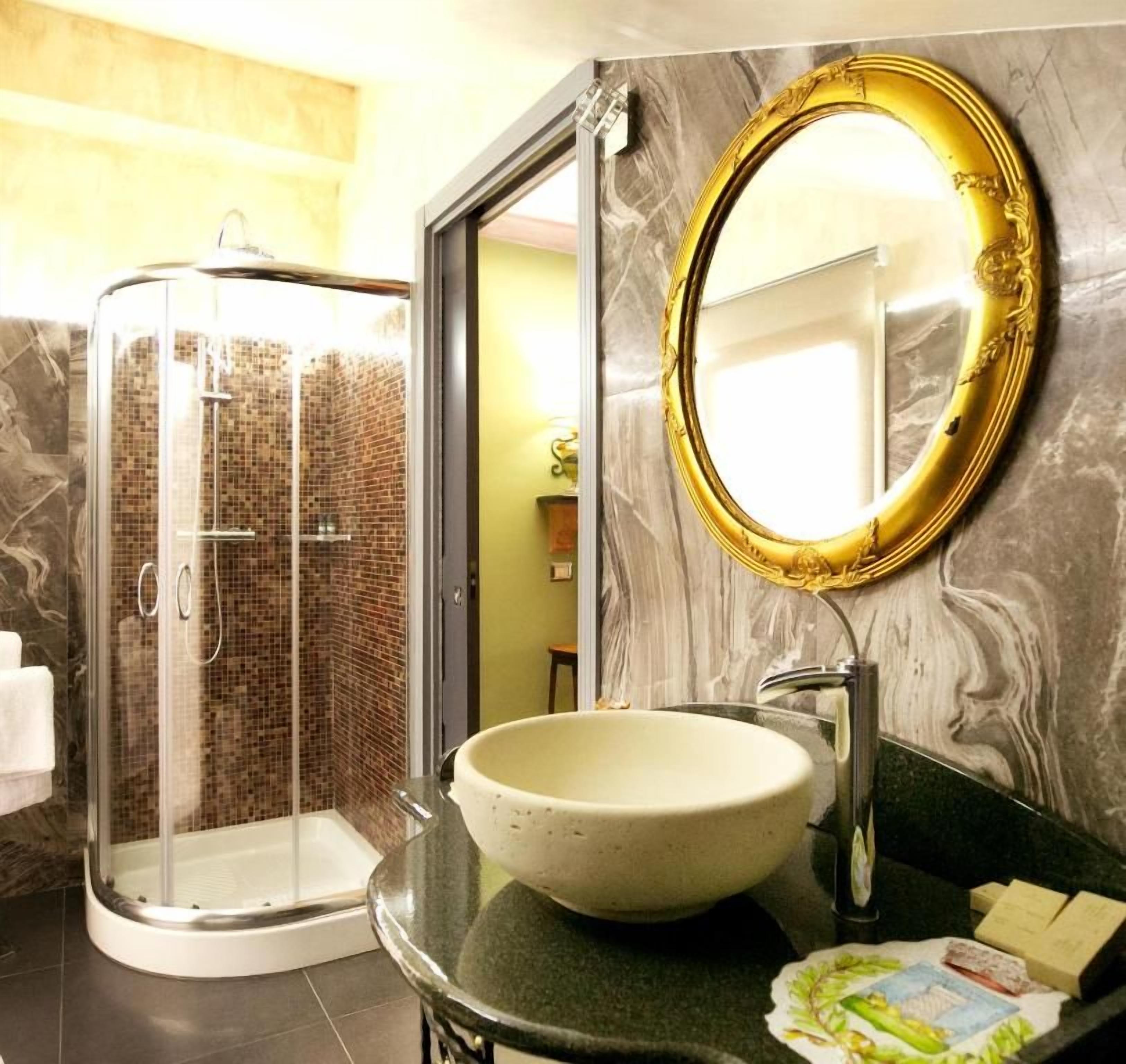 Un bagno con doccia vetrata, parete in marmo, specchio con cornice dorata e lavandino bianco.