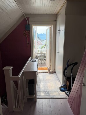 Un pasillo estrecho con piso de madera, barandilla blanca en un lado y una puerta que da a un balcón con vistas a casas y montañas.