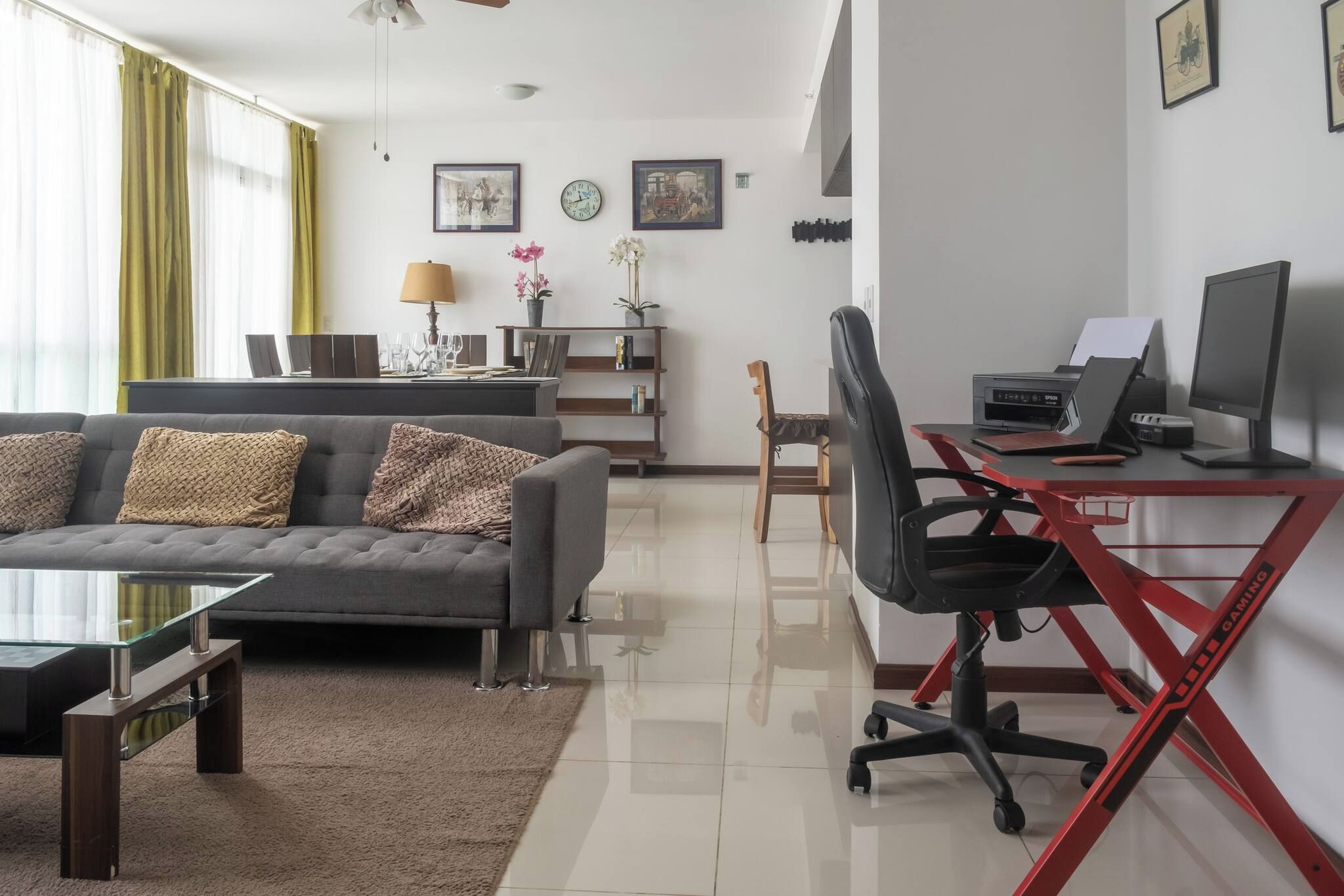 A modern living room with a grey sofa, a glass coffee table, and a red gaming desk with a computer and monitor.