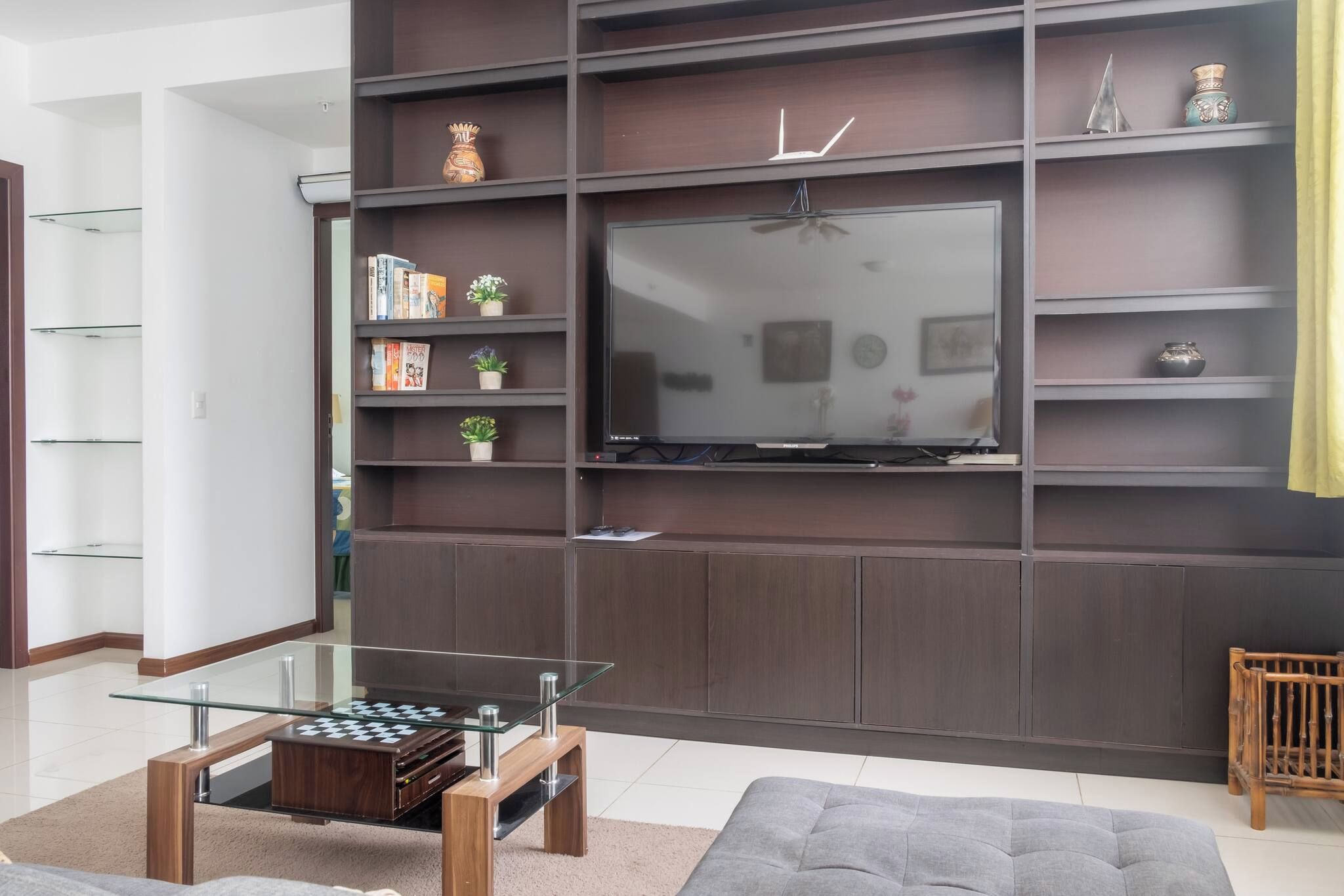 A modern living room with a flat-screen TV mounted on a dark wood panel wall, a glass coffee table with a chess set, and a grey sofa.