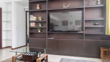 A modern living room with a flat-screen TV mounted on a dark wood panel wall, a glass coffee table with a chess set, and a grey sofa.