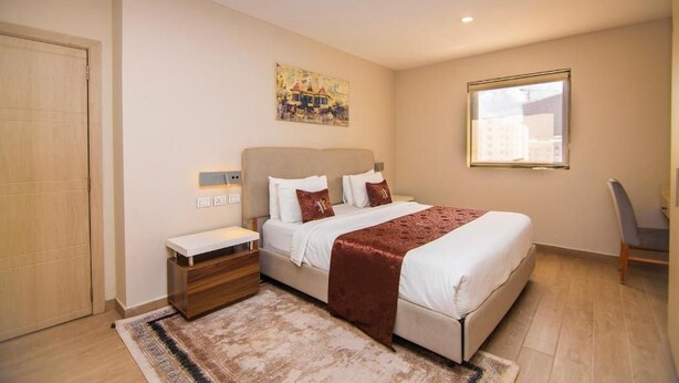 Un dormitorio con cama, mesitas de noche, una silla, una ventana y un cuadro en la pared.