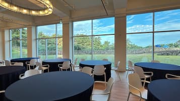 A spacious indoor area with round tables and chairs, large windows offering a view of a garden and sky.