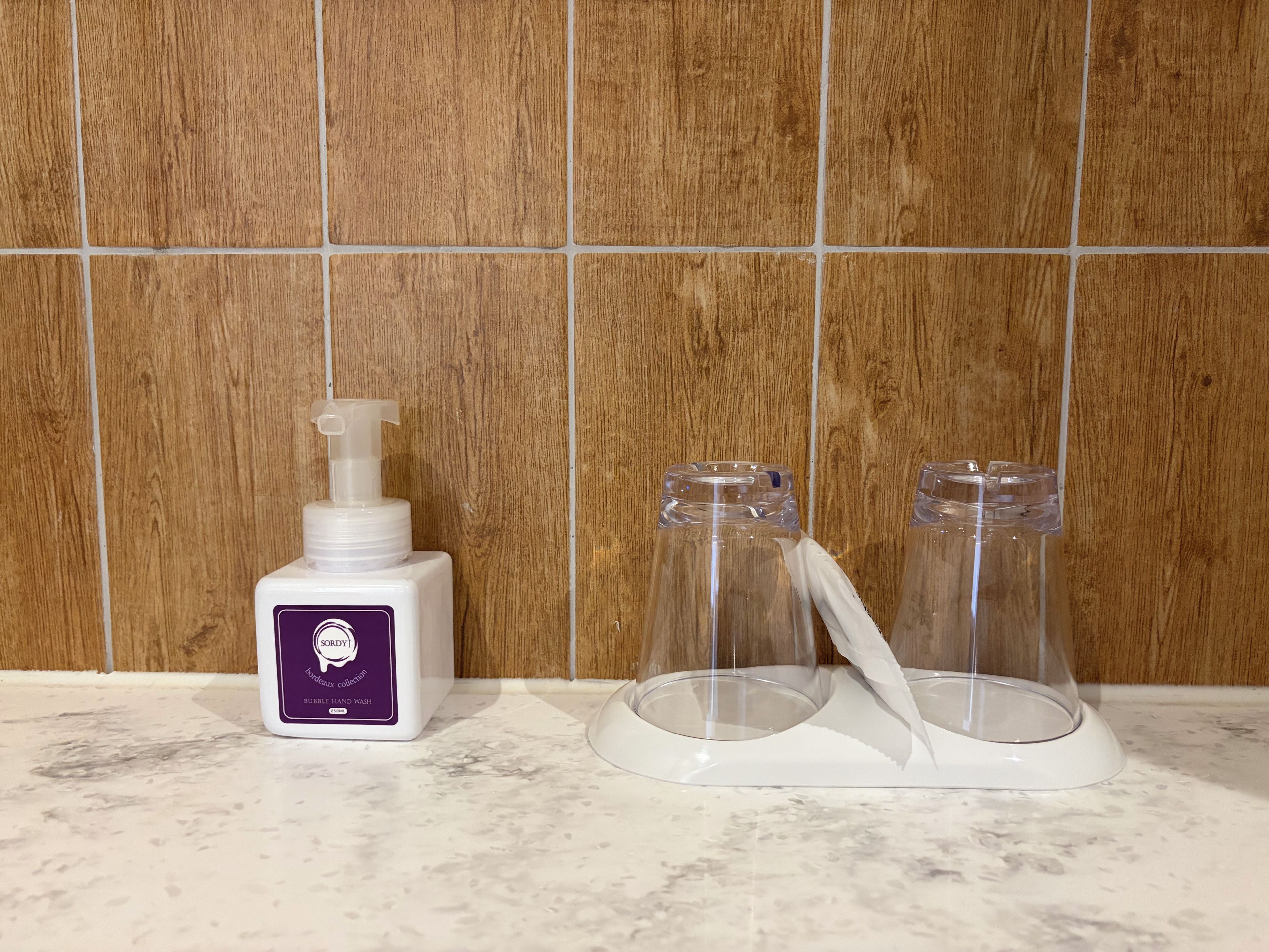 Un baño con pared de madera, una botella de jabón para manos y dos recipientes de vidrio sobre una encimera de mármol.