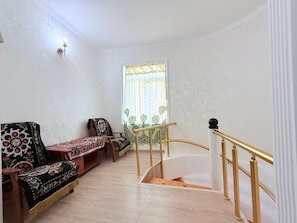 Uma sala de estar com um sofá, uma escada com corrimão dourado e uma janela com cortinas estampadas.