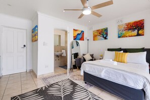 Un dormitorio con cama, ventilador de techo, baño con lavamanos y una puerta.