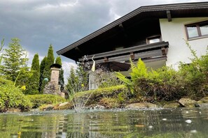 A modern house with a stone monument, a pond, and lush greenery. - Alpine garden in the holiday home Weimar (Ellmau)