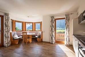 Ein Zimmer mit einem Holztisch, einer Bank und Blick ins Freie durch große Fenster.