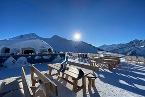 Ein Außenbereich mit Holzbänken und Tischen, im Hintergrund ein mit Schnee bedeckter Berg und ein klarer Himmel.