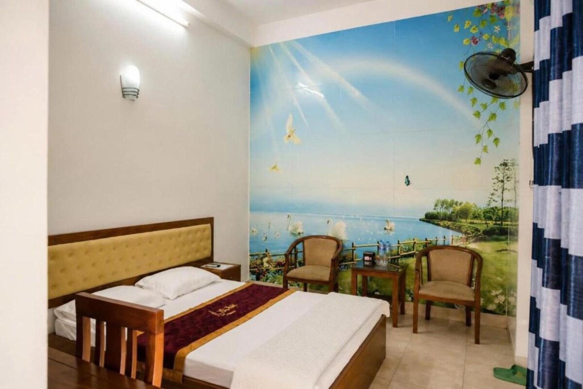 A hotel room with a bed, two chairs, a small table, and a mural of a landscape with birds.