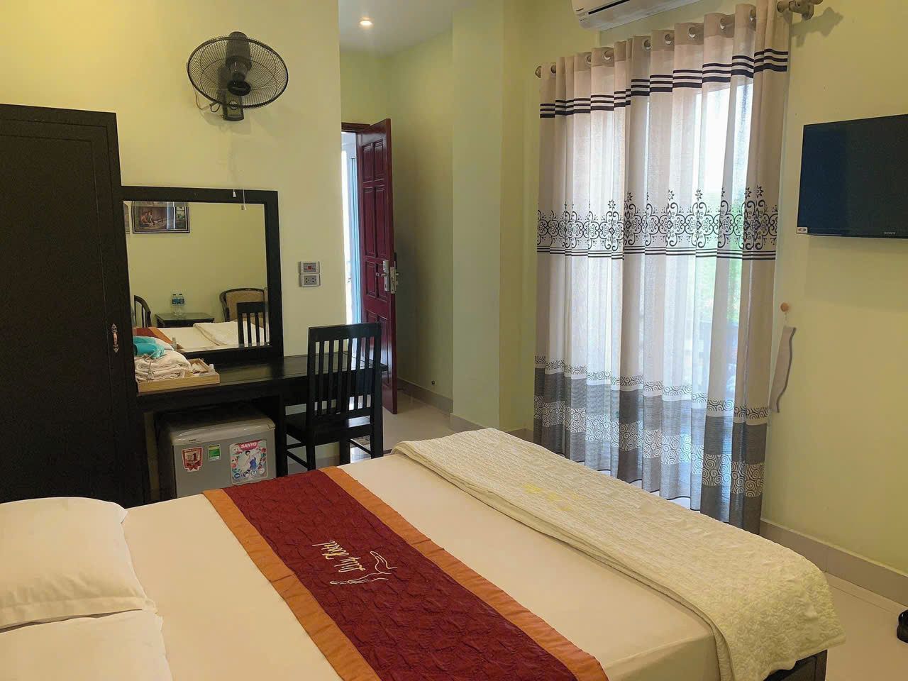 A hotel room with a bed, a desk, a chair, a mirror, a fan, and curtains.