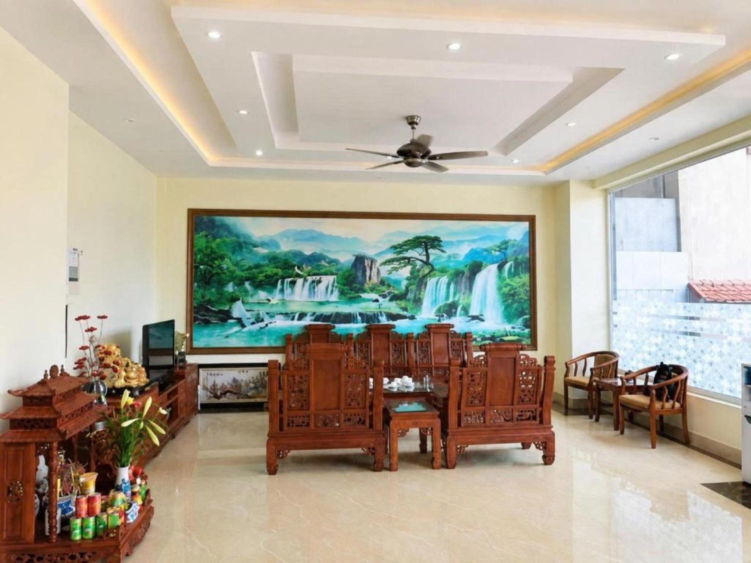 Un salón amplio con ventilador de techo, un cuadro grande de una cascada, muebles de madera tradicionales y suelo de baldosas.