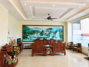 A spacious living area with a ceiling fan, a large painting of a waterfall, traditional wooden furniture, and a tiled floor.