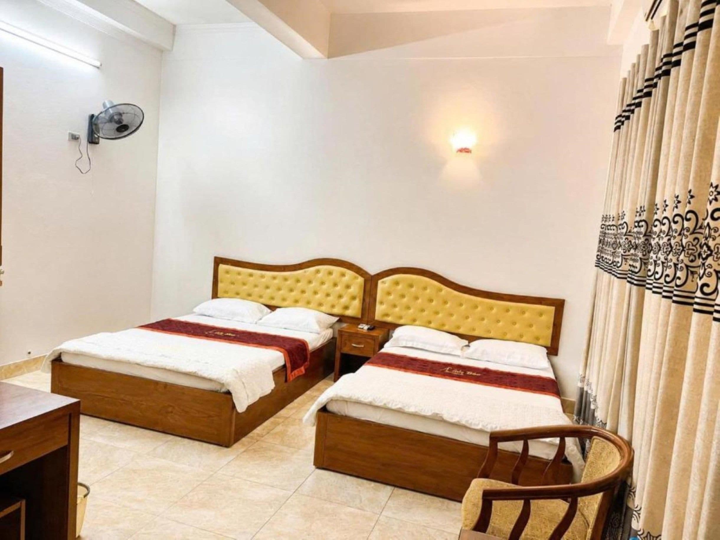 Quarto de hotel com duas camas, uma mesinha de cabeceira, uma cadeira, um ventilador fixado na parede e uma cortina estampada.