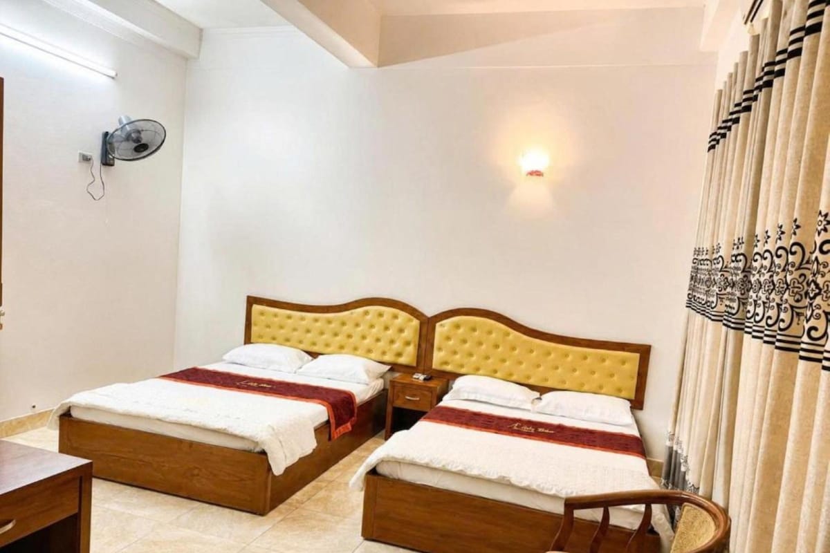 A hotel room with two beds, a bedside table, a chair, a wall-mounted fan, and a curtain with a pattern.