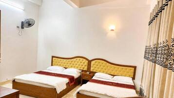 Quarto de hotel com duas camas, uma mesinha de cabeceira, uma cadeira, um ventilador fixado na parede e uma cortina estampada.