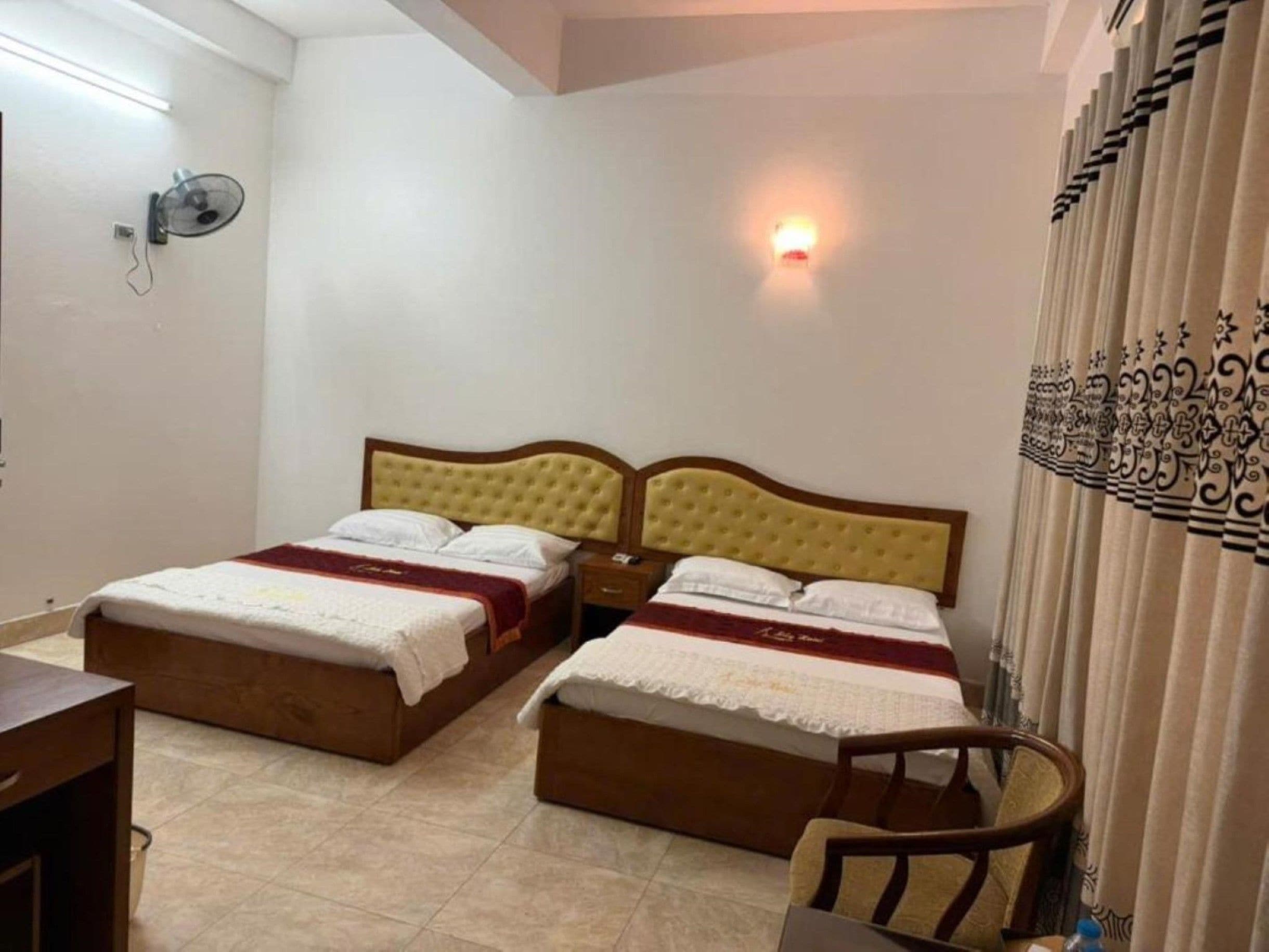 A hotel room with two beds, a bedside table, a chair, and a wall-mounted fan.