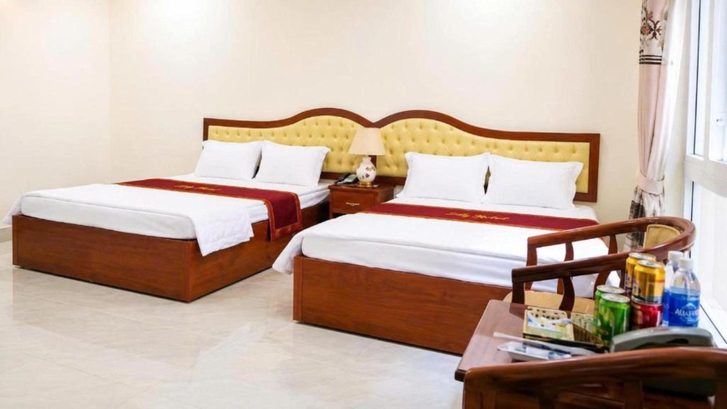 A hotel room with two beds, a wooden headboard, a nightstand, a lamp, and a small table with a chair.