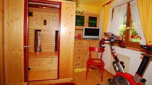A room with a wooden cabinet, a red chair, a red exercise bike, and a window with curtains. - Maple view in the Tasser house (Mayrhofen)