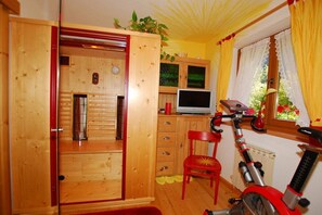 A room with a wooden cabinet, a red chair, a red exercise bike, and a window with curtains. - Maple view in the Tasser house (Mayrhofen)