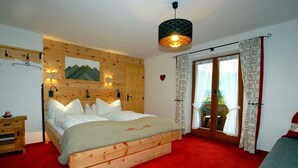 A bedroom with a wooden bed, a red carpet, a wooden wall panel, a window with curtains, and a hanging light fixture. - Maple view in the Tasser house (Mayrhofen)