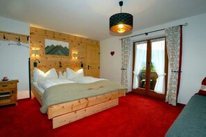 A bedroom with a wooden bed, a red carpet, a wooden wall panel, a window with curtains, and a hanging light fixture. - Maple view in the Tasser house (Mayrhofen)