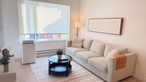 Un salon avec un canapé, une table basse, une lampe et une fenêtre avec des stores.