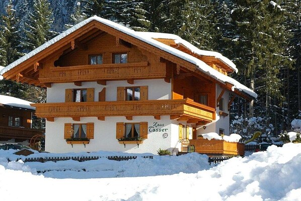A two-story wooden chalet with a snow-covered roof, surrounded by trees and a snow-covered ground. - Penkenblick in the Tasser House (Mayrhofen)