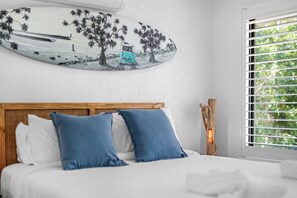 A bed with white bedding and blue pillows, a wooden headboard, a wall-mounted artwork depicting a car and trees, and a window with blinds. - Jindiburra Coastal Haven at Noosaville (Noosaville)