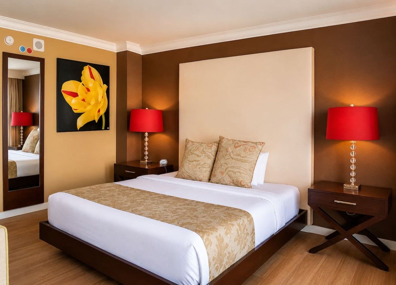 Habitación de hotel con una cama grande, dos mesitas de noche con lámparas, un espejo y un cuadro en la pared.