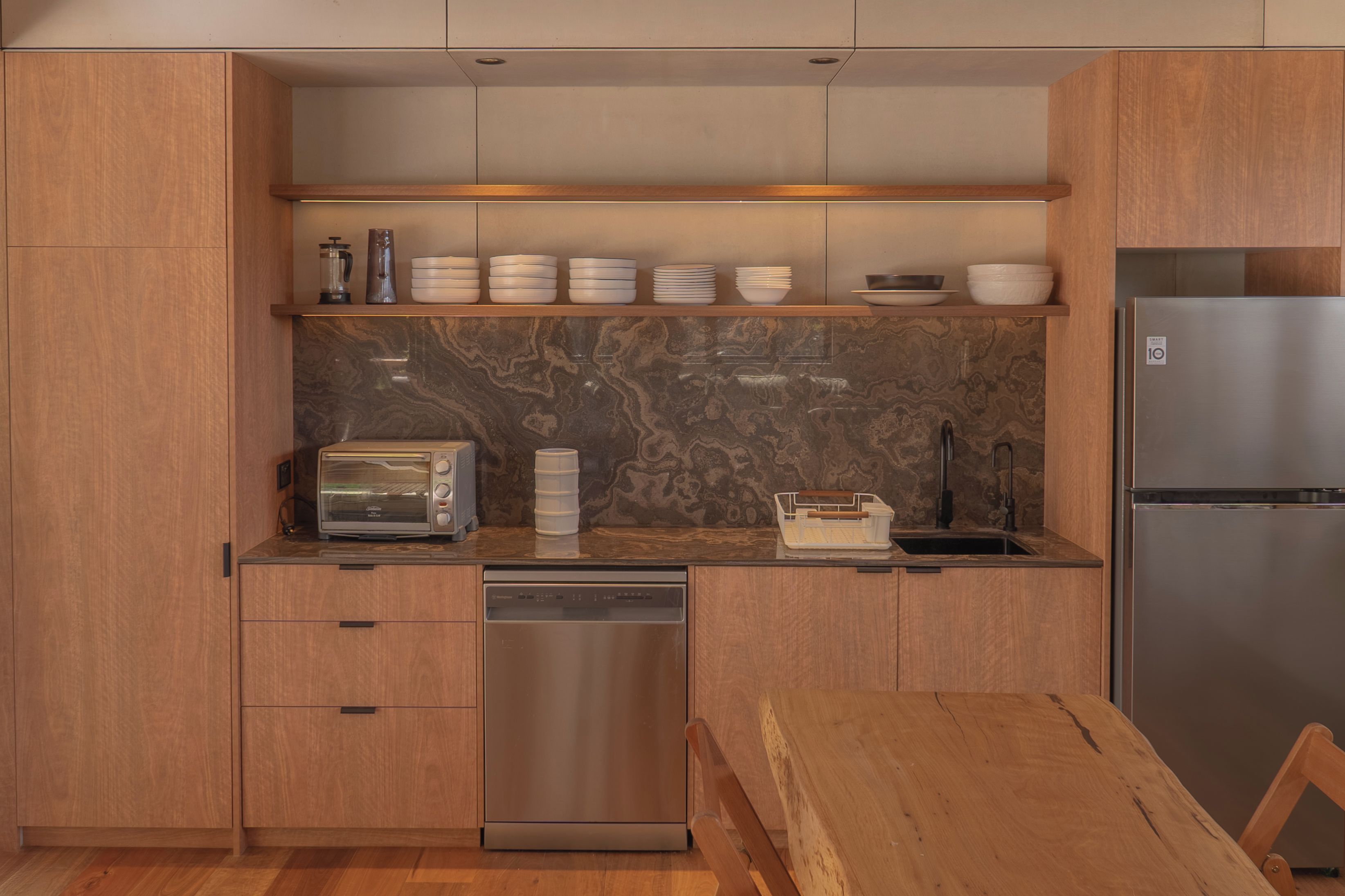 A modern kitchen with wooden cabinets, a built-in dishwasher, a stainless steel refrigerator, and a marble backsplash.