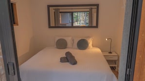 Uma cama com colcha branca, duas almofadas e uma toalha enrolada. Tem uma mesa de cabeceira com abajur e uma janela acima da cama.