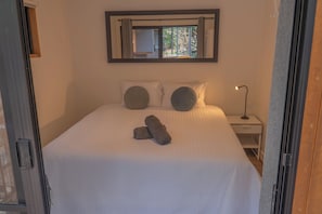 A bed with a white bedspread, two pillows, and a rolled towel. There is a nightstand with a lamp and a window above the bed. - Private Hinterland Cabin near Beaches & Rail Trail: Cabin 2 (Burringbar)