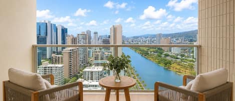 A balcony with two wicker chairs, a small round table with a vase, and a view of a cityscape and river.