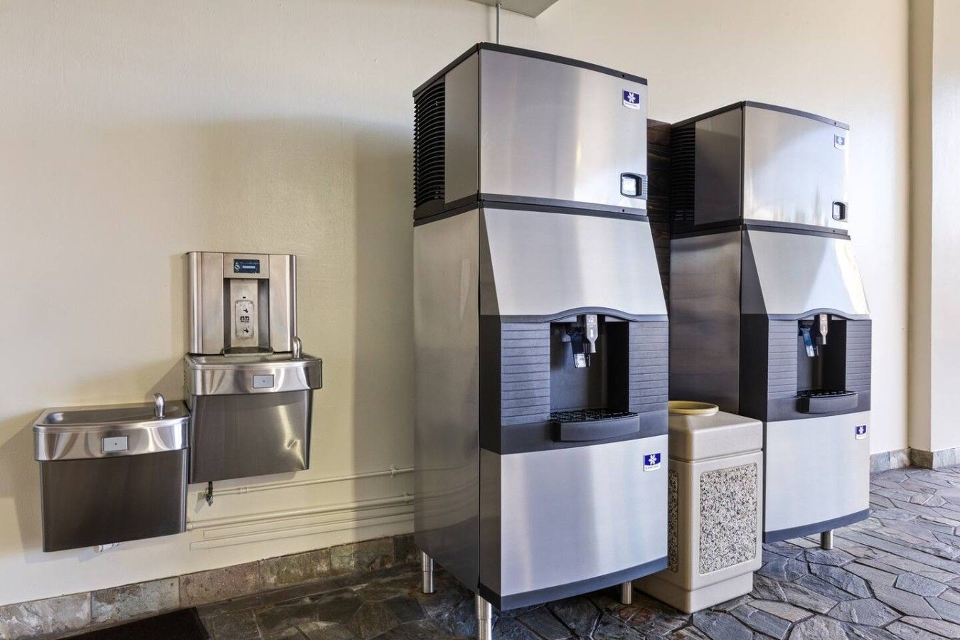 A water dispenser and trash bin mounted on a wall, with a stone-tiled floor.