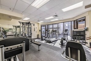 Un gimnasio con diversos equipos de ejercicio, amplias ventanas y piso alfombrado.
