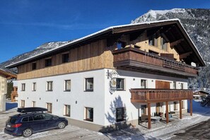 Ein zweistöckiges Gebäude mit Balkon, ein Auto davor und verschneite Berge im Hintergrund.