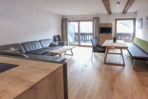 A modern living room with a sofa, a dining table, and a TV mounted on the wall. - 60 m² Guesthouse Sonne (Bichlbach)