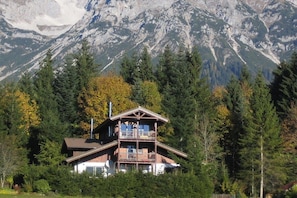 A two-story wooden cabin with a balcony, surrounded by dense forest and mountains in the background.