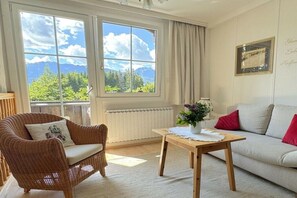 A room with a view of mountains, a wicker chair, a wooden table, and a white sofa with red cushions.