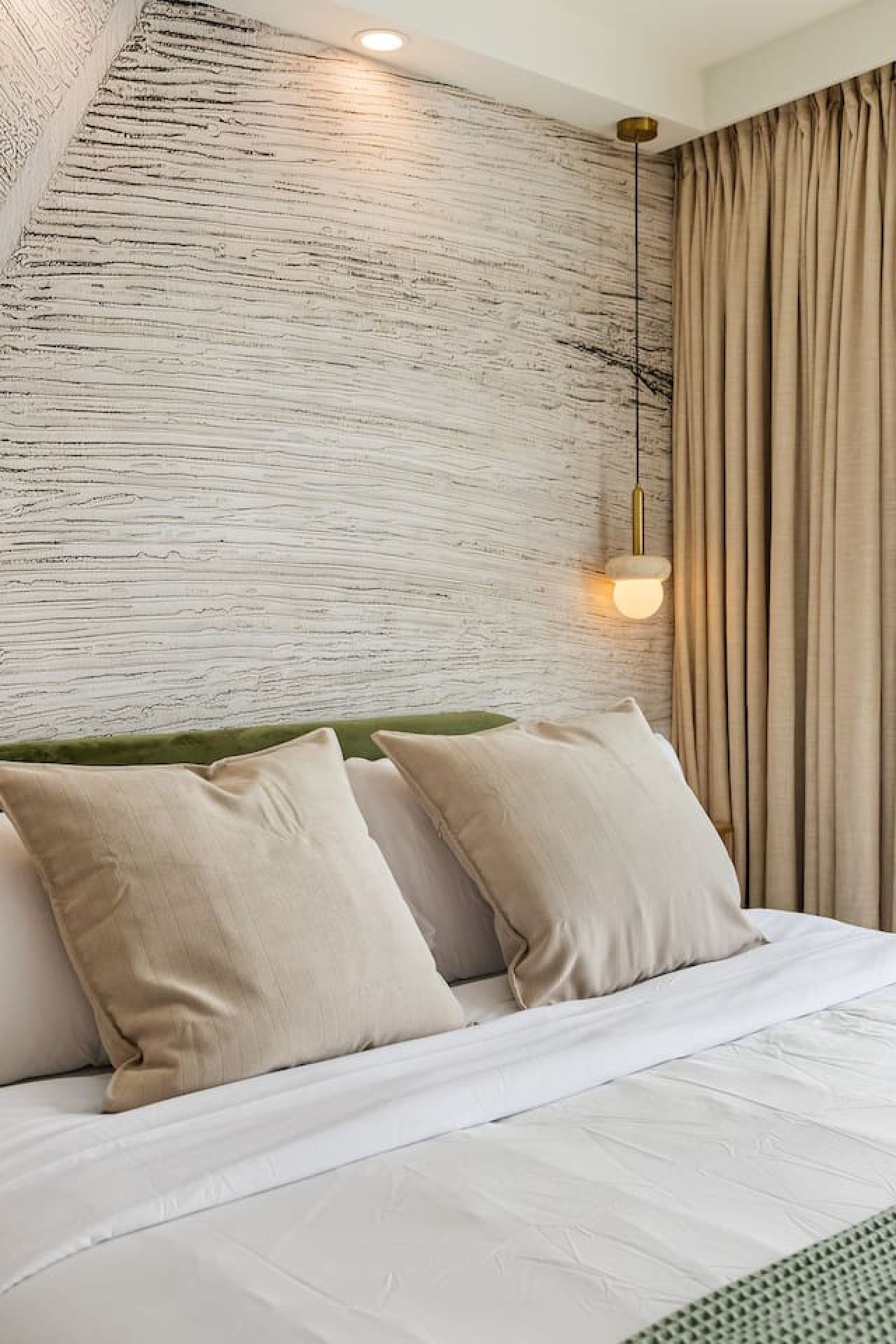 A modern bedroom with a textured wall, a bed with white bedding, and a hanging light fixture.