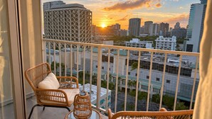 A balcony with wicker furniture, a view of a cityscape at sunset, and a building with a grid-like facade. - New Waikiki Studio | King | Parking | Pool (Honolulu)